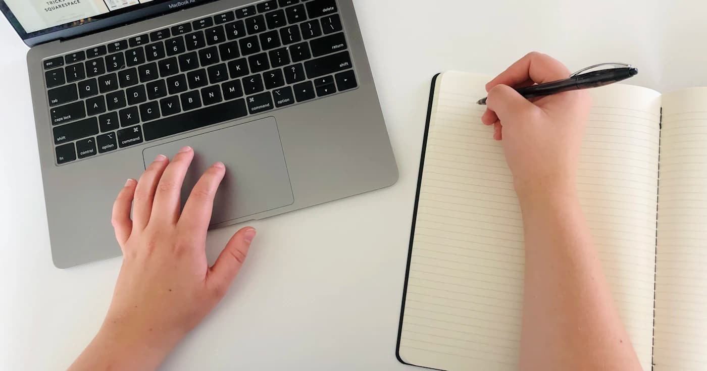 Person writing gift ideas in a notebook beside a laptop